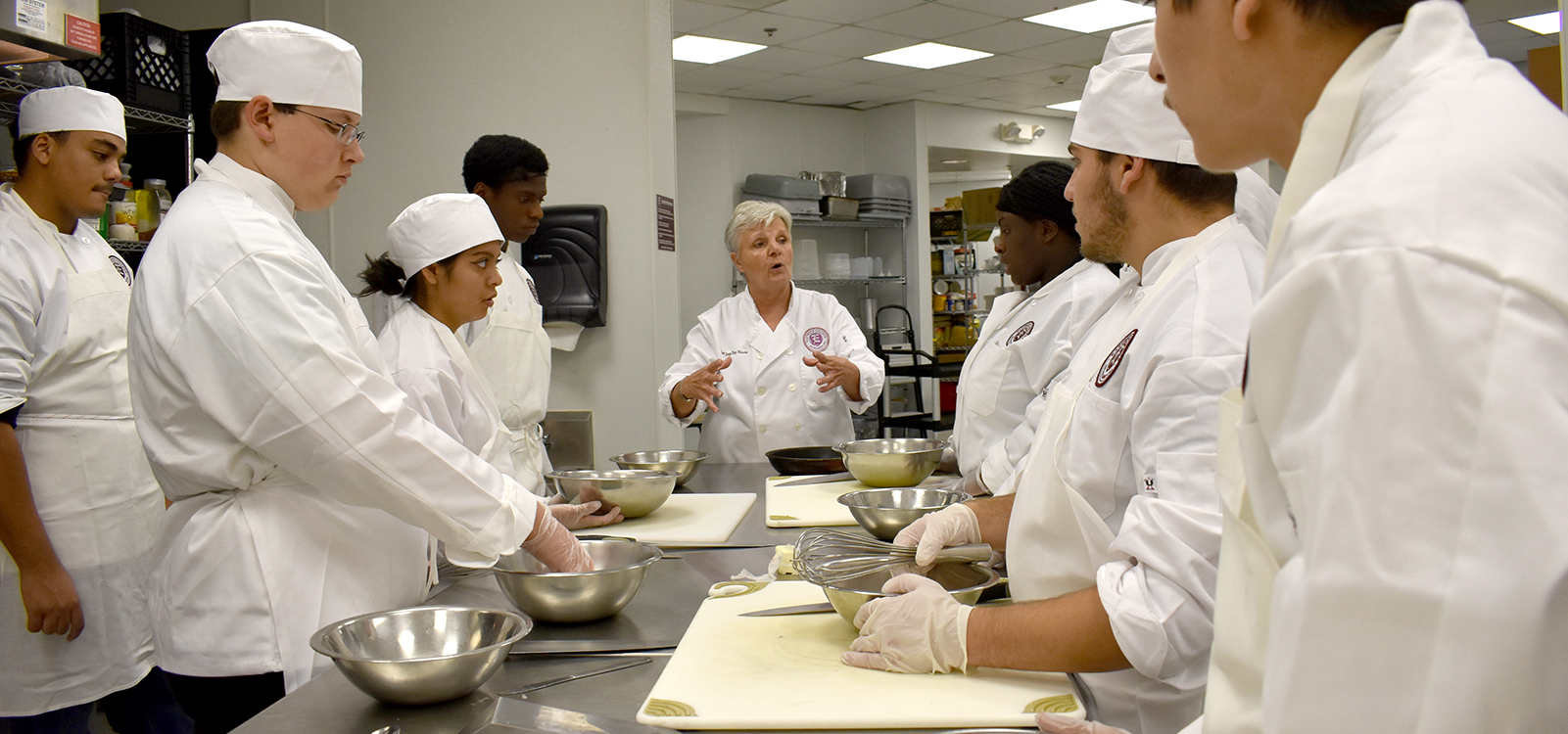 Culinary Arts Associates students stand around table full of kitchen tools listening to instructor
