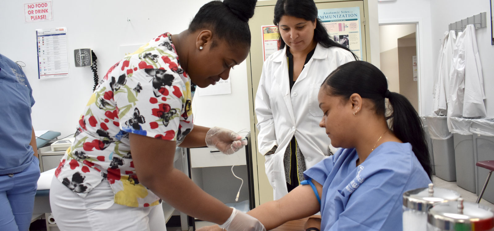 medical assisting student practices drawing blood on fellow student while instructor watches